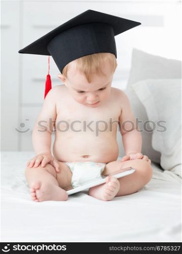 10 months old baby boy in diapers wearing graduation cap and using tablet pc