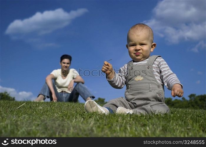 10 months old baby is playing outdoor while his mother is relaxing in the background.