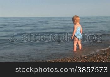 20 month old girl stands on a pebble shore and looks out to sea