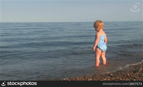 20 month old girl stands on a pebble shore and looks out to sea