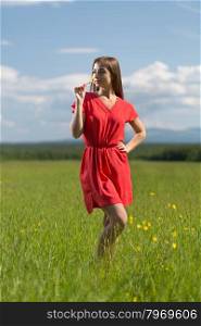 20 year-old girl in red dress sniffing a yellow flower in a field on a sunny day