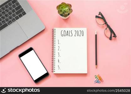 2020 New year concept. Goals list in stationery, laptop, notebook, smartphone, pot plant on pink pastel color with copy space