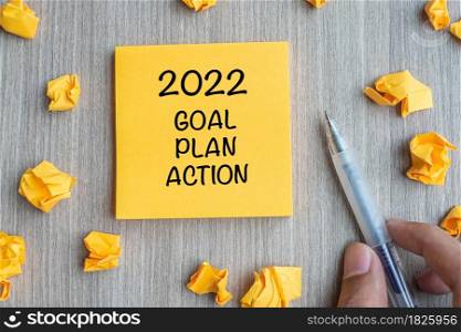 2022 Goal, Plan, Action word on yellow note with Businessman holding pen and crumbled paper on wooden table background. New Year New Start, Resolutions, Strategy concept