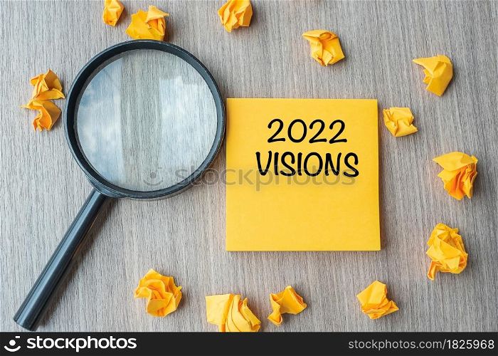 2022 VISIONS words on yellow note with crumbled paper and magnifying glass on wooden table background. New Year New Start, Idea, Strategy, and Goals concept