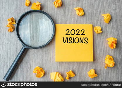 2022 VISIONS words on yellow note with crumbled paper and magnifying glass on wooden table background. New Year New Start, Idea, Strategy, and Goals concept