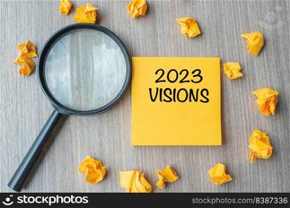 2023 VISIONS words on yellow note with crumbled paper and magnifying glass on wooden table background. New Year New Start, Idea, Strategy, and Goals concept