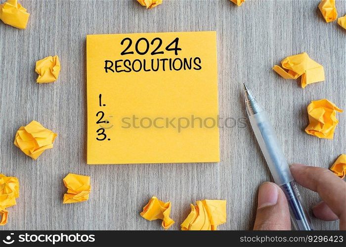2024 RESOLUTIONS word on yellow note with Businessman holding pen and crumbled paper on wooden table background. New Year start, Strategy and Goal concept