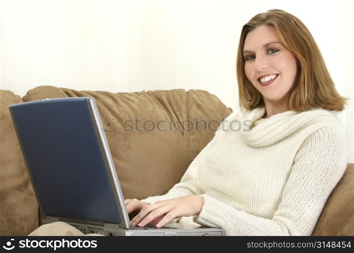 25 year old woman on the couch working on a laptop computer. Wearing cream sweater, smiling.
