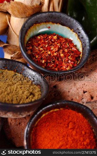 3 dark rimmed glazed ceramic bowls filled with cumin, crushed red pepper & red chile powder; displayed upon a porous rock with ladder in the background.