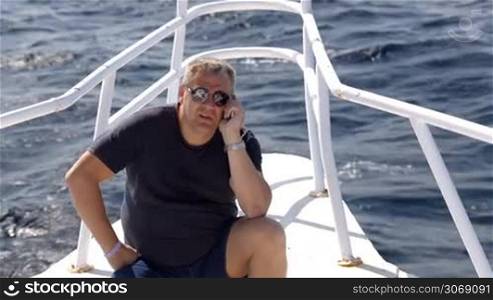 50 years old man sitting on the bow of sailing yacht and talking on the phone