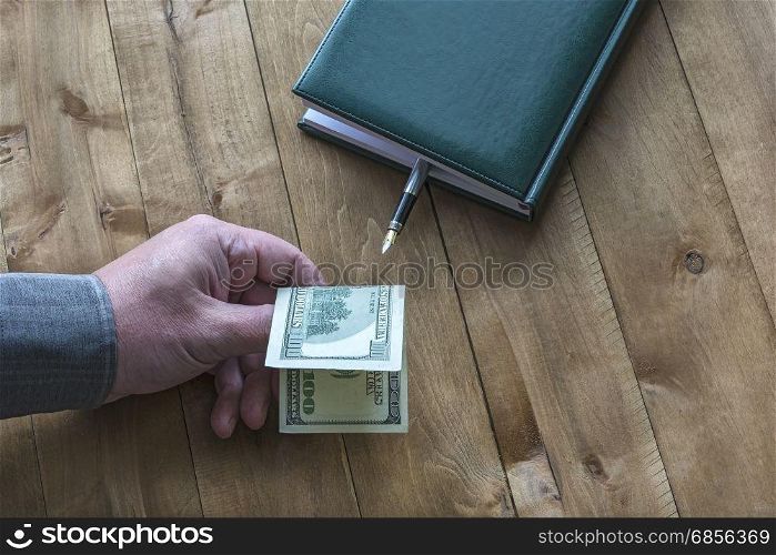A $ 100 dollar bill is held by a man&rsquo;s hand near a diary with a fountain pen on a wooden surface.