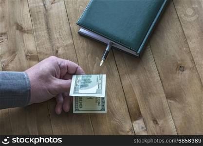 A $ 100 dollar bill is held by a man&rsquo;s hand near a diary with a fountain pen on a wooden surface.