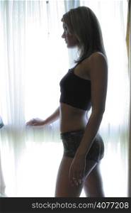 a 20&acute;s woman dressed in lingerie stands at window silhouetted.