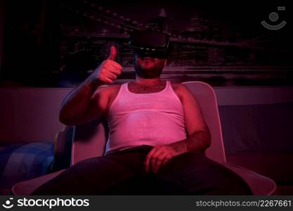 A 40-year-old unshaven overweight man watches TV with vr glasses, shows various emotions, approves . lazy middle-aged man