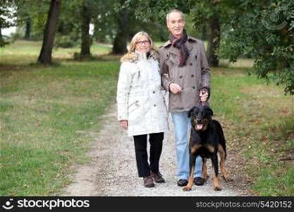 a 60 years old woman holding husband&rsquo;s arm in a park in autumn, the man is keeping a dog on the leash