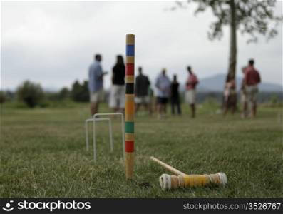 A backyard croquet game in progress. The mallet and end stake are in focus with the players blurred with depth of field.