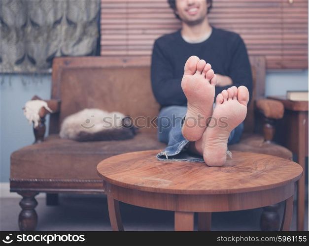 A barefoot young man is resting his legs on a coffee table at home, there is a cat on the sofa next to him