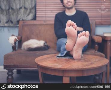 A barefoot young man is resting his legs on a coffee table at home, there is a cat on the sofa next to him