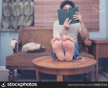 A barefoot young man is resting his legs on a coffee table at home while reading, there is a cat on the sofa next to him