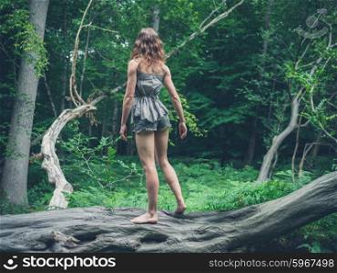 A barefoot young woman is standing on a fallen tree in the forest surrounded by ferns