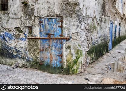 A battered old blue door with a bar for security in a run-down neighborhood is located at a street corner in the Mellah portion of Essaouira in Morocco.