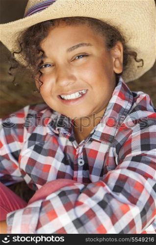 A beautiful and happy mixed race African American female girl child wearing straw cowboy hat and plaid shirt sitting in hay filled barn