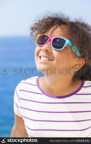 A beautiful and happy mixed race African American female girl child wearing sunglasses in bright sunshine on vacation in front of a blue tropical sea