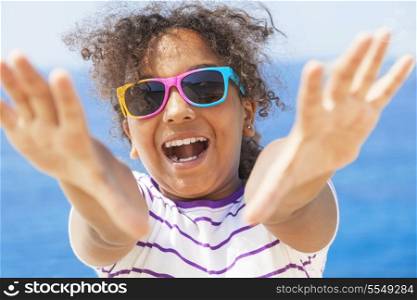 A beautiful and happy mixed race African American female girl child wearing sunglasses in bright sunshine on vacation in front of a blue tropical sea laughing &amp; reaching to camera