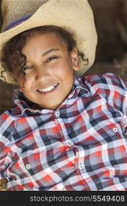 A beautiful and happy mixed race African American female girl child wearing straw cowboy hat and plaid shirt sitting in hay filled barn