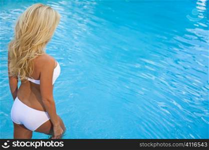A beautiful and sexy blond haired woman wearing a white bikini walking into a turquoise blue spa pool, sea or ocean lit by natural golden evening sunshine