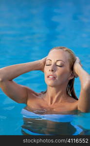 A beautiful and sexy young blond woman wearing a white bikini pushes her hair back as she emerges from a turquoise blue swimming pool. Spa, healthy living and health club concept.