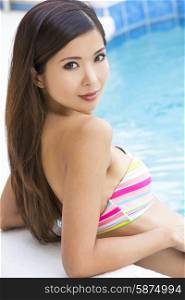 A beautiful and sexy young chinese asian woman wearing a bikini leaning on side of a turquoise blue swimming pool. Spa, healthy living and health club concept.