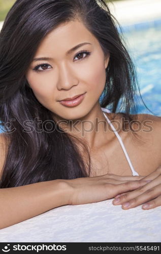 A beautiful and sexy young Chinese Asian woman wearing a white bikini leaning on side of a turquoise blue swimming pool. Spa, healthy living and health club concept.