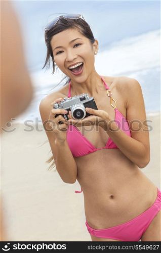 A beautiful Asian Chinese girl or young woman wearing pink bikini at a beach looking happy taking pictures or photographs with a retro digital camera