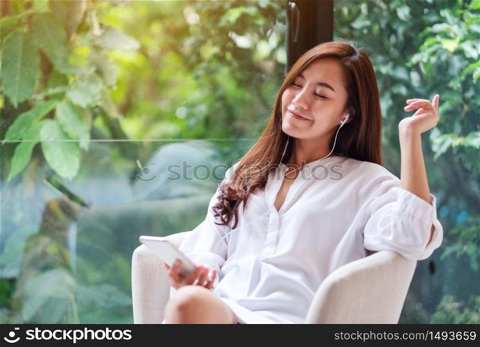 A beautiful asian woman enjoy listening to music with phone and earphone at home , green nature background , happiness and relaxation concepts
