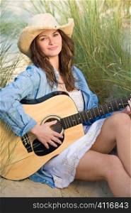 A beautiful blond brunette woman sitting in amongst the sand dunes and tall grass illuminated by natural late evening golden sunshine having fun playing her guitar