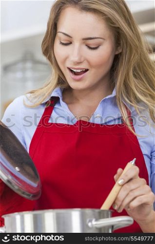 A beautiful blond girl or young woman looking happy wearing a red apron &amp; cooking in her kitchen at home