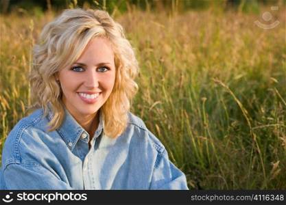 A beautiful blond haired blue eyed model sits amid tall grass illuminated by golden evening sunlight