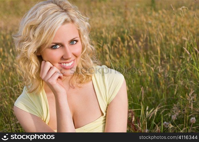 A beautiful blond haired blue eyed model sits amid tall grass illuminated by golden sunlight