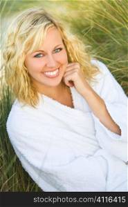A beautiful blond haired blue eyed model wearing a white towelling robe sits amid tall grass
