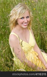 A beautiful blond haired blue eyed model wearing a yellow dress sits amid tall grass.