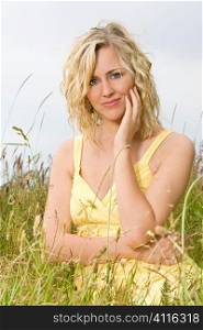 A beautiful blond haired blue eyed model wearing a yellow dress sits amid tall grass