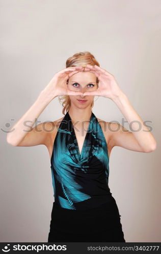 A beautiful blond woman holding her hands before her eyes and makinga sign, like a photographer, what the picture will look like, over light graybackground.