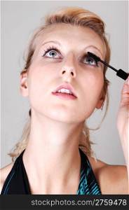 A beautiful blond woman in closeup, doing her makeup and fixing hereyelashes, looking up, for light gray background.