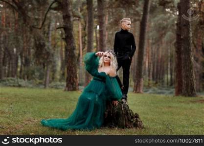 A beautiful blonde young woman in a long green dress and a diadem on her head with stylish young son in the forest. girl and boy sitting near the old stump. Solar glare. Fantasy. fairy tale. A beautiful blonde young woman in a long green dress and a diadem on her head with stylish young son in the forest. girl and boy sitting near the old stump. Solar glare. Fantasy. fairy tale.