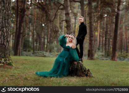 A beautiful blonde young woman in a long green dress and a diadem on her head with stylish young son in the forest. girl and boy sitting near the old stump. Solar glare. Fantasy. fairy tale. A beautiful blonde young woman in a long green dress and a diadem on her head with stylish young son in the forest. girl and boy sitting near the old stump. Solar glare. Fantasy. fairy tale.
