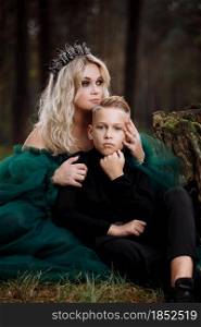 A beautiful blonde young woman in a long green dress and a diadem on her head with stylish young son in the forest. girl and boy sitting near the old stump. Solar glare. Fantasy. fairy tale.. A beautiful blonde young woman in a long green dress and a diadem on her head with stylish young son in the forest. girl and boy sitting near the old stump. Solar glare. Fantasy. fairy tale
