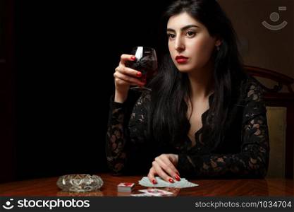 a beautiful brunette woman with scarlet lips in black clothes is sitting at a table, drinking wine and playing cards for undressing