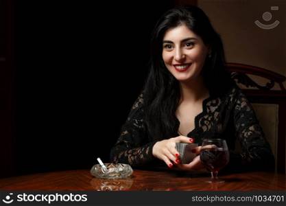 a beautiful brunette woman with scarlet lips in black clothes sits at a table and plays cards for undressing