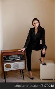 a beautiful Caucasian young woman in a black pantsuit and black sandals stands next to a vintage record player